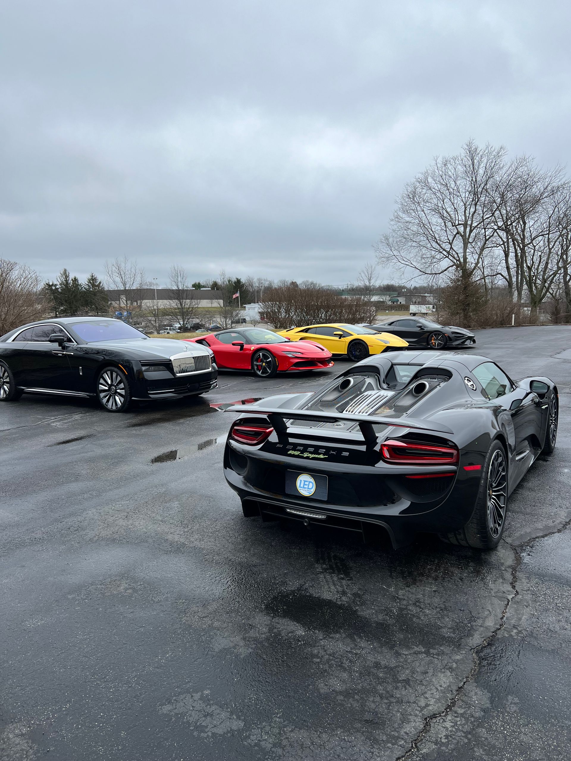 Several luxury and sports cars, including a black Porsche 918 Spyder and a Rolls-Royce, parked on an overcast, wet lot.