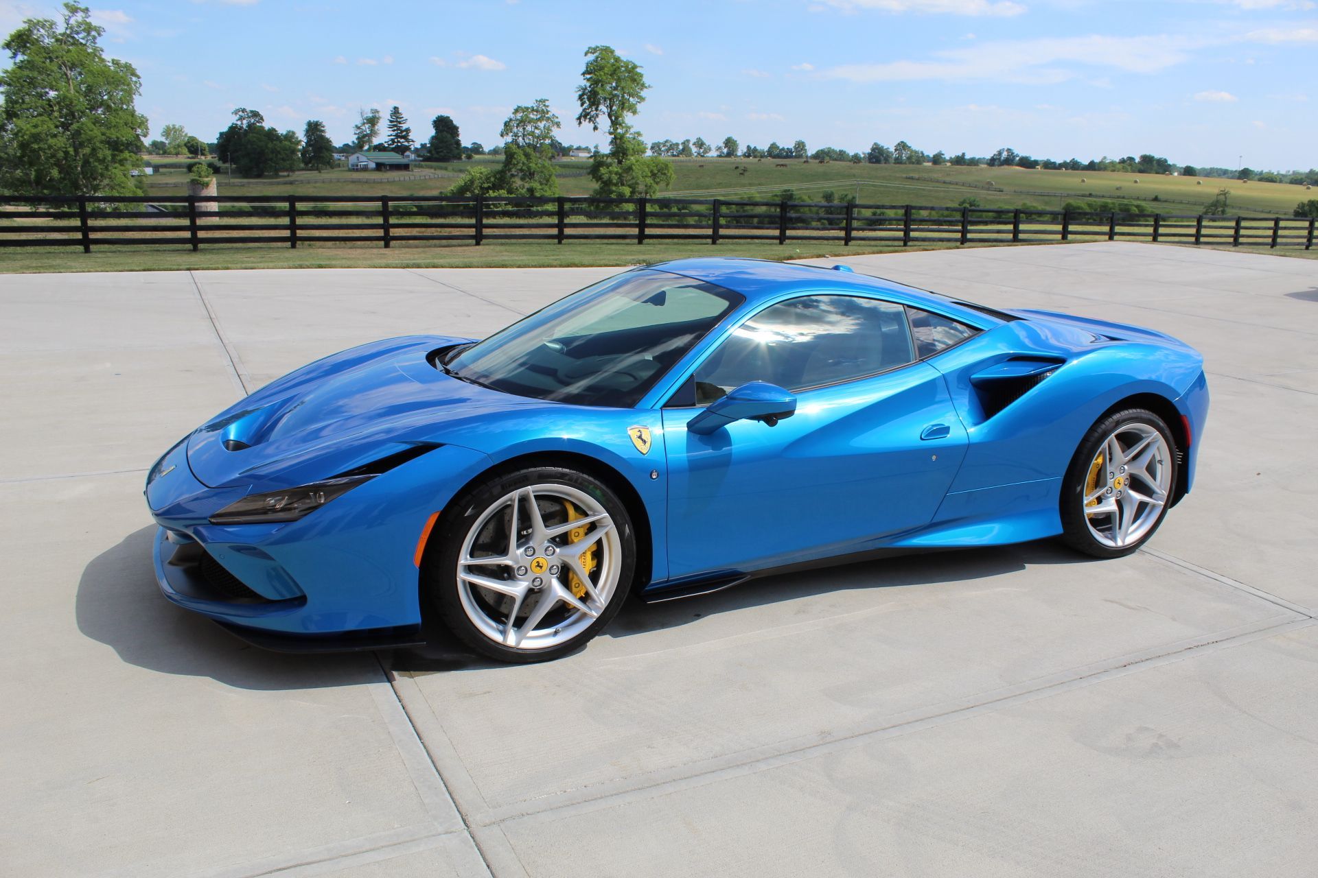 A bright blue Ferrari F8 Tributo parked on a concrete lot against a backdrop of a field and a wooden fence.