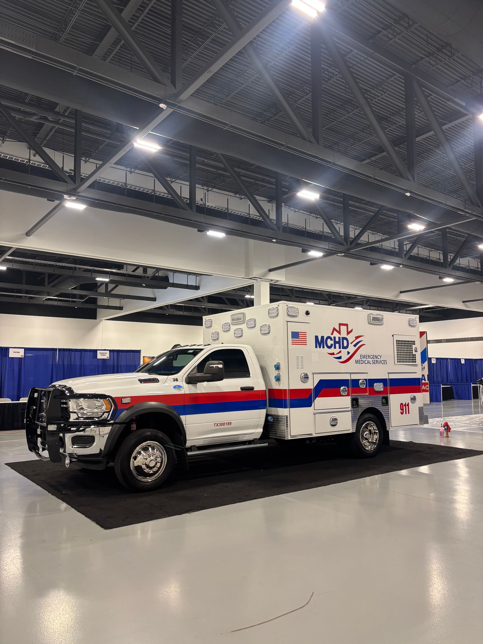 A white and blue MCHD ambulance parked on a black floor in a large, brightly lit indoor hall.