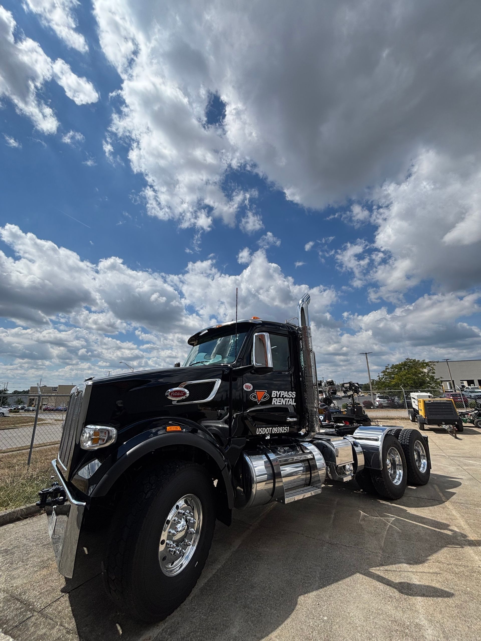 A black semi-truck with chrome accents parked on a gravel lot under a bright, cloudy blue sky.