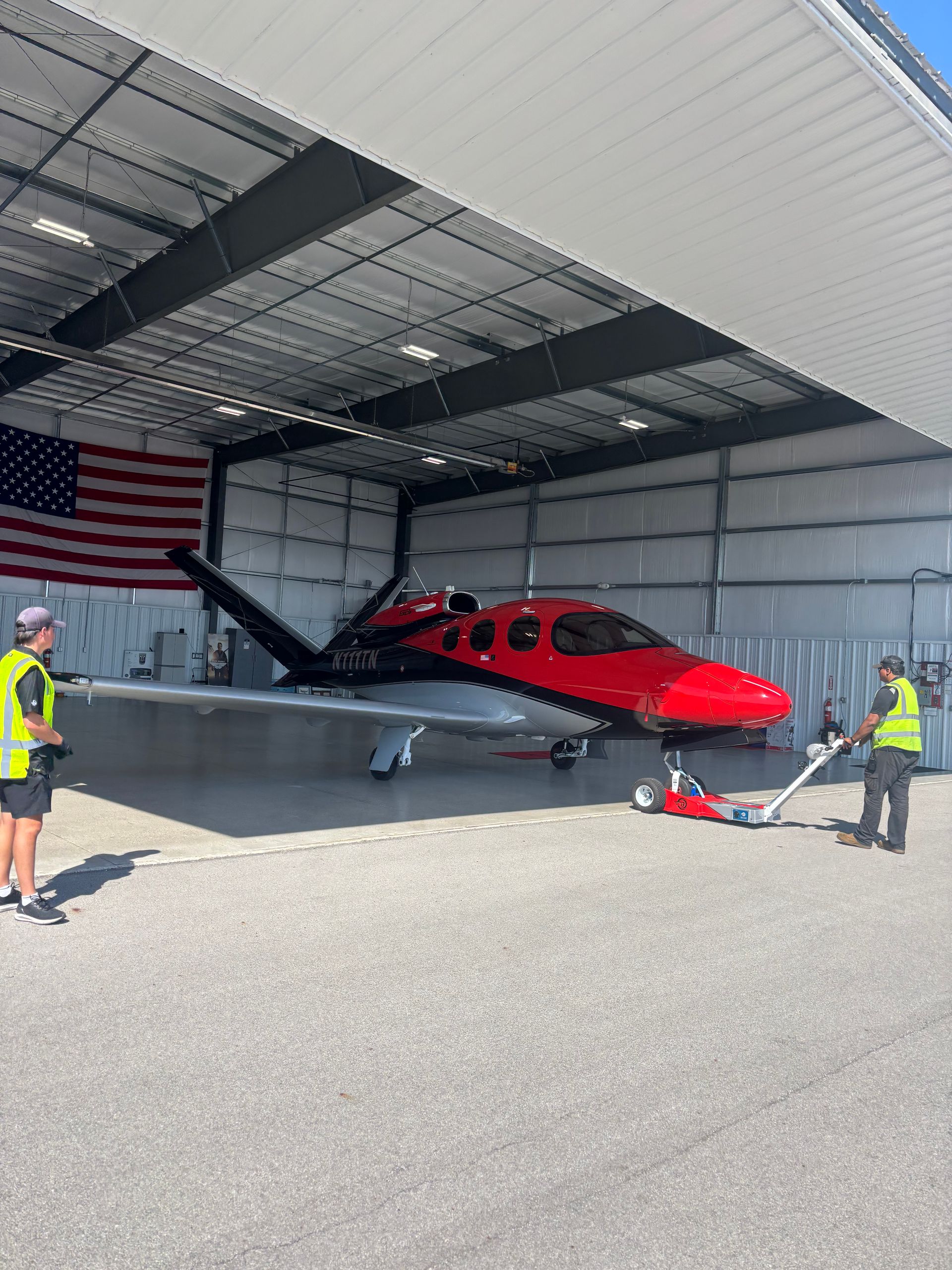 Two people use a tow tug to move a red and silver Cirrus Vision Jet out of a hangar featuring an American flag.