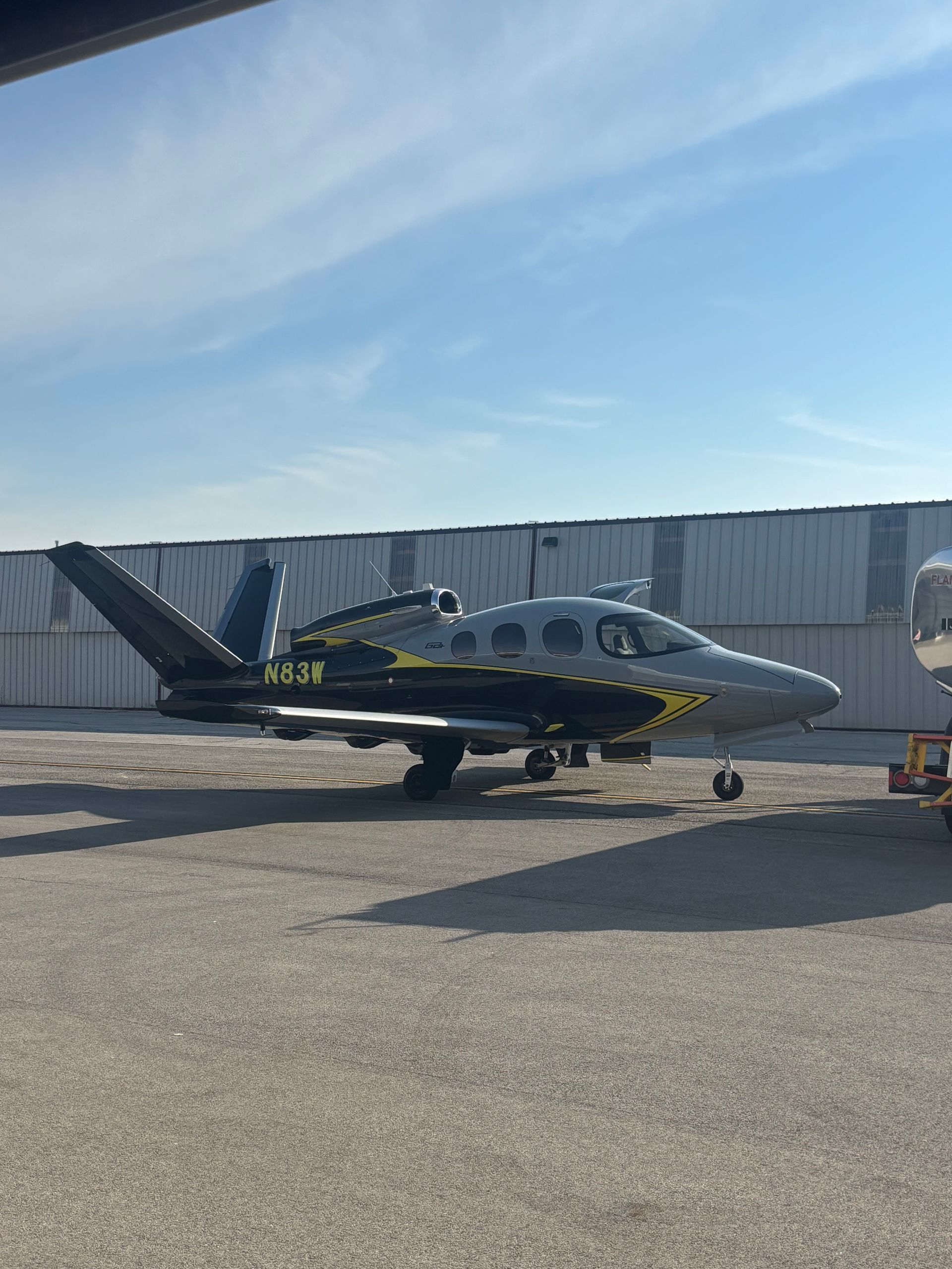 A parked silver and black business jet with a V-tail design on a gravel ramp in front of a hangar under a blue sky.