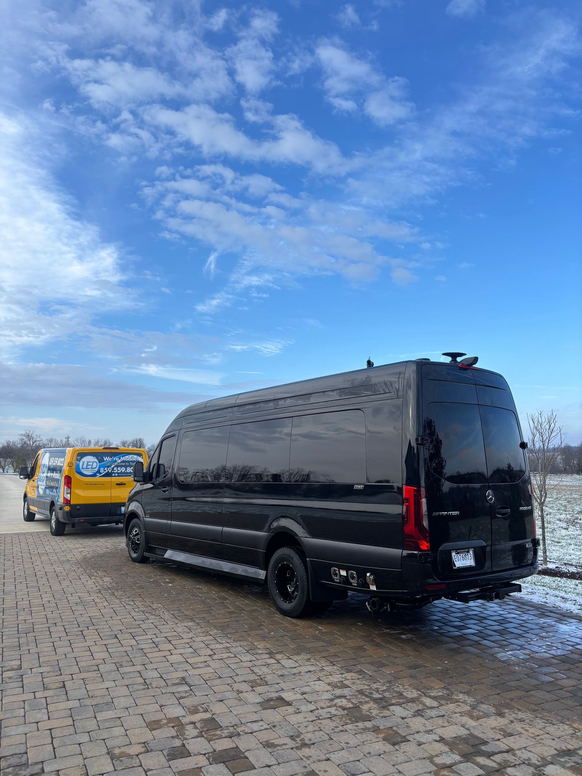 A large black customized cargo van parked on a cobblestone path next to a yellow utility van on a snowy, sunny day.