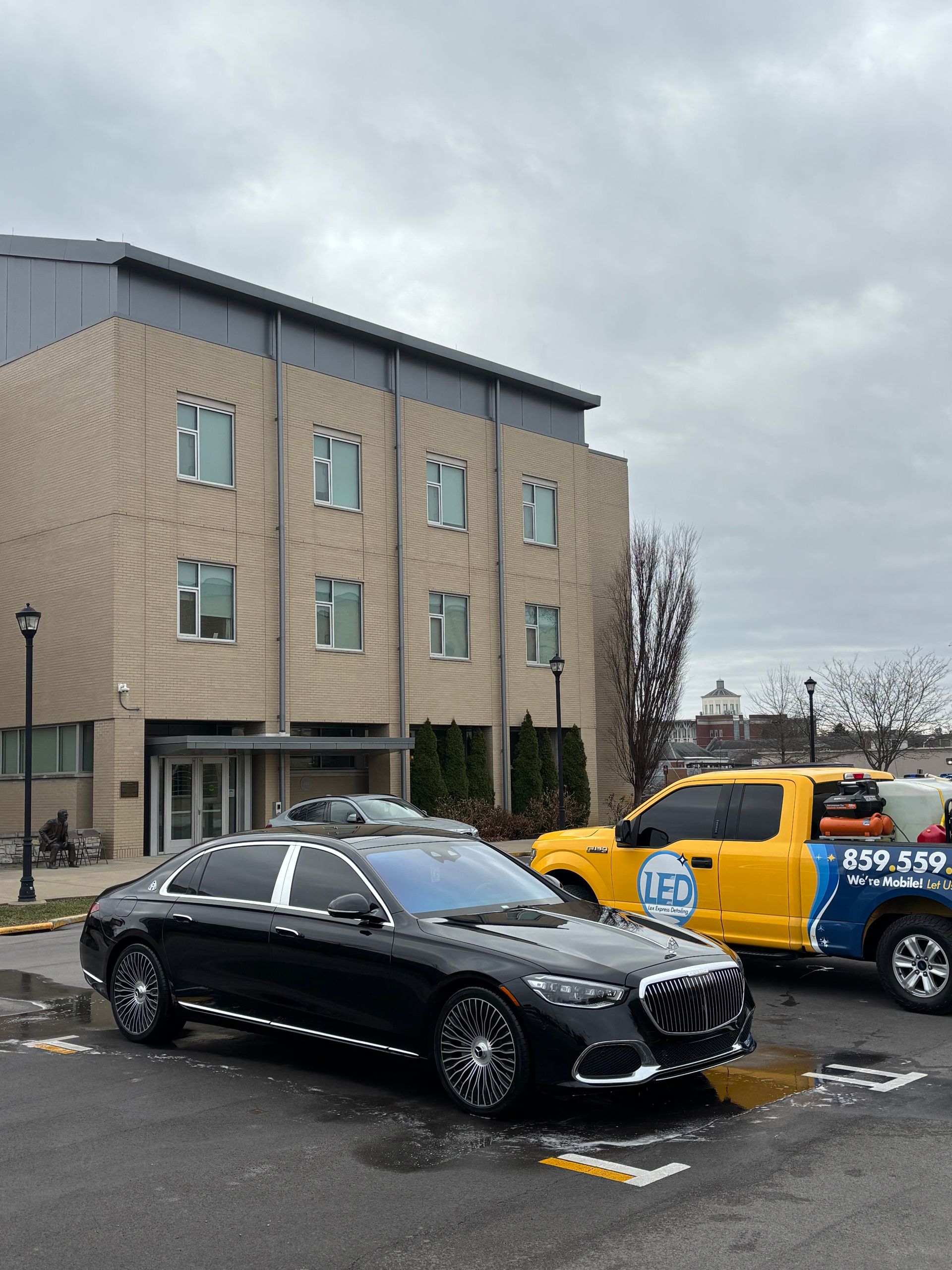 A black luxury sedan is parked in a parking lot next to a yellow utility truck in front of a multi-story brick building.