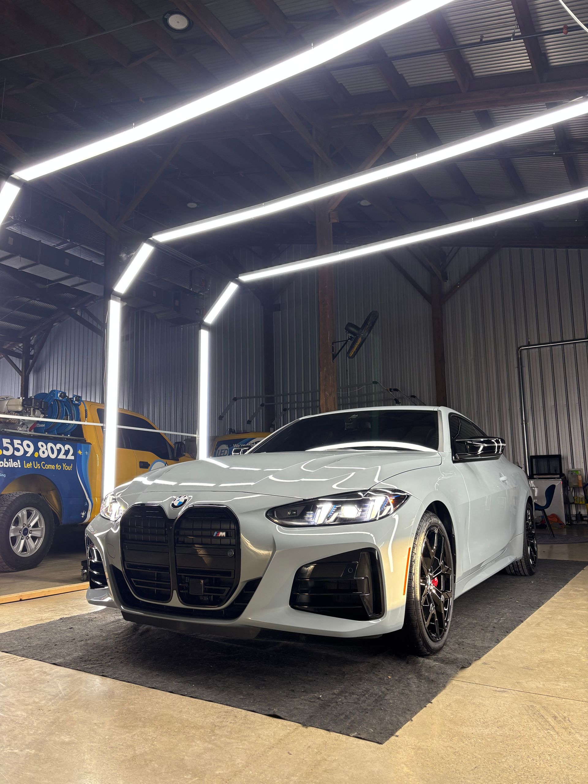 A gray BMW M4 parked inside a garage lit by arched overhead LED light frames.