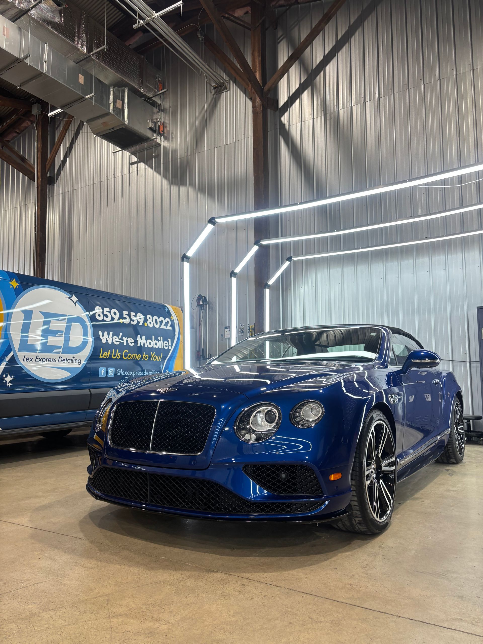 A deep blue Bentley parked inside a garage lit by arched overhead LED lights.