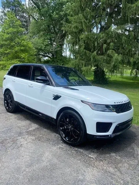 A white Range Rover Sport with black wheels and a black roof parked on an asphalt driveway near green trees.