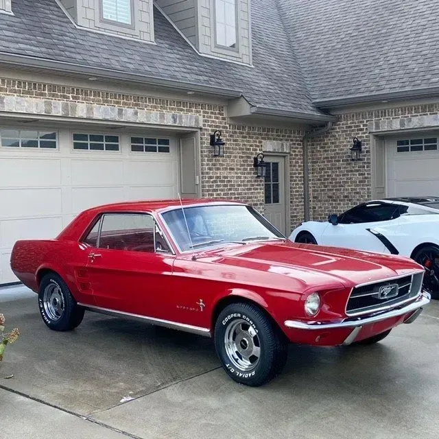 A classic red Ford Mustang parked on a residential driveway in front of a brick house with a white sports car nearby.