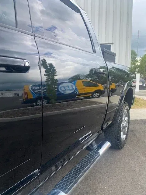 A side view of a black pickup truck with a yellow service van reflected in its glossy door panel.