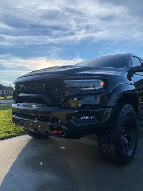 A black RAM 1500 TRX pickup truck parked on a driveway against a bright, cloudy sky.