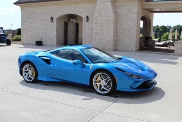 A bright blue Ferrari F8 Tributo parked on a paved driveway in front of a stone building.