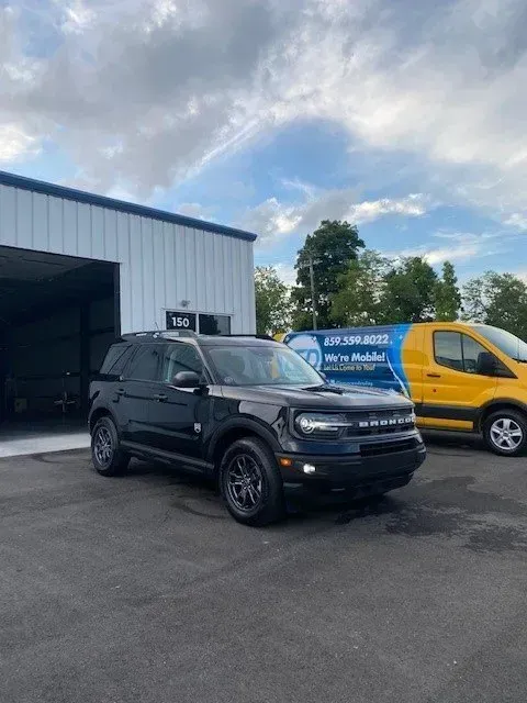 A black Ford Bronco Sport parked in a paved lot in front of a white building, next to a yellow commercial van.