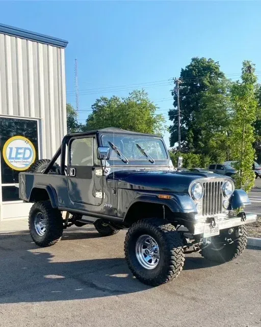 A dark blue vintage Jeep with large off-road tires parked outside a building with an LED sign.
