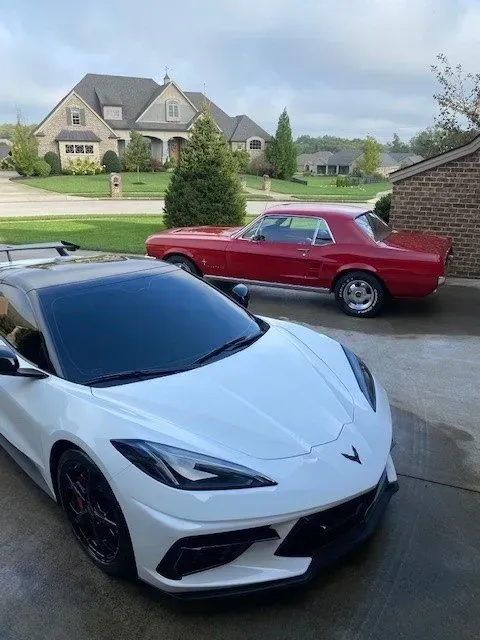 A white Corvette and a red vintage muscle car parked in a residential driveway in front of a suburban home.