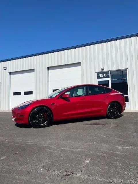A red Tesla parked in front of a commercial warehouse building with white metal siding on a sunny day.