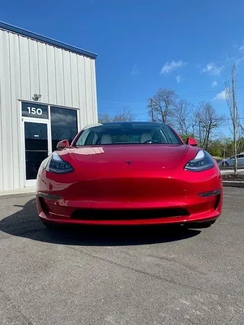 A glossy red Tesla Model 3 parked in front of a white building with the number 150 on the glass door.