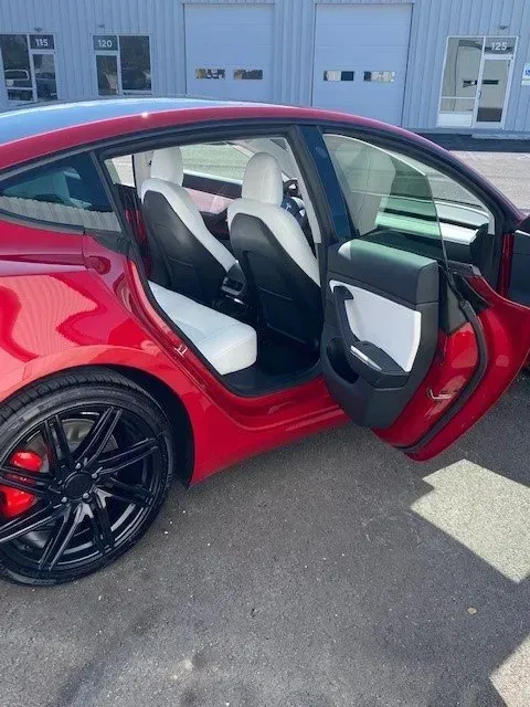 A red Tesla Model 3 with black rims and red brake calipers parked outdoors, with the rear passenger door open.