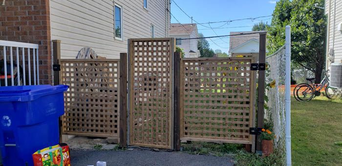 A wooden lattice fence with a matching gate between a house and a chain-link fence, with a blue bin in the foreground.