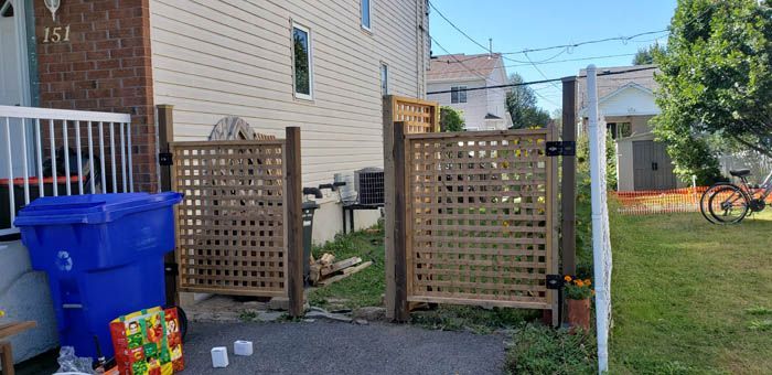 Two wooden lattice gates stand side-by-side in a driveway next to a tan house, with a blue recycling bin in the foreground.