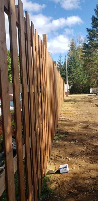 A close-up, angled view of a newly installed wooden picket fence running along a dirt path under a bright, cloudy sky.