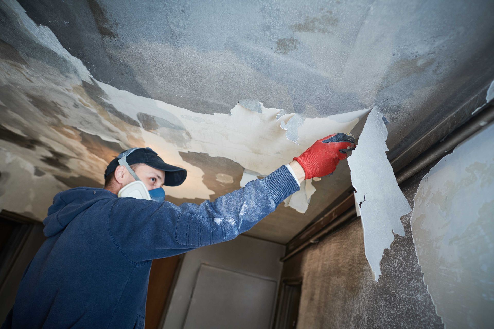 A worker wearing a respirator and red gloves peels old, damaged wallpaper off a ceiling in a room under renovation.