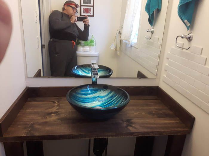 A person taking a selfie in a bathroom with a rustic wooden vanity and a circular blue and teal glass vessel sink.
