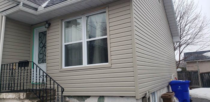 Tan vinyl siding house exterior with a white-framed window, a front door, and a black metal railing on the porch steps.