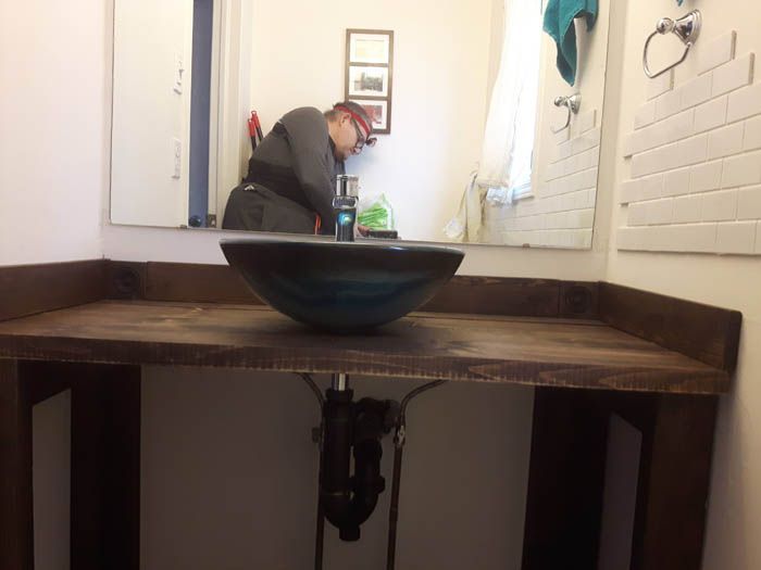 A dark wood bathroom vanity with a bowl-style sink, viewed from the front, with a person reflected in the mirror behind it.