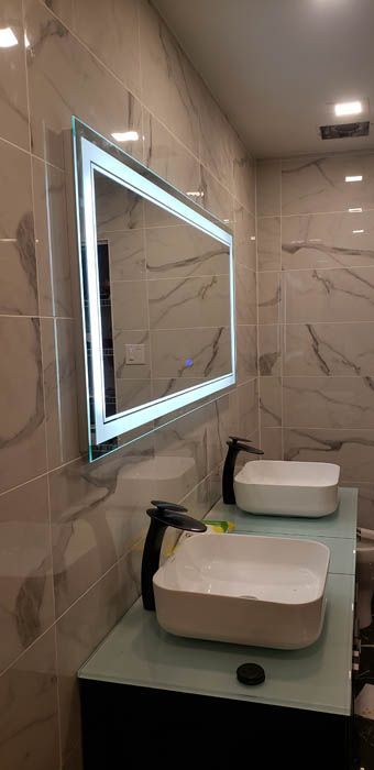 A modern bathroom vanity with dual square white sinks, black faucets, and a rectangular LED-lit mirror on marble walls.