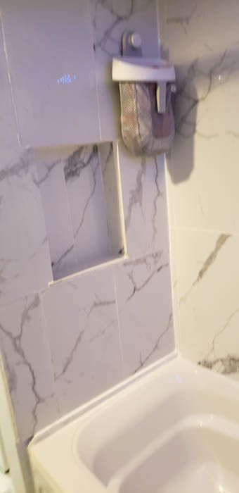 A shower wall with white marble-patterned tiles, a recessed shelf, and a mounted soap dispenser above a white basin.