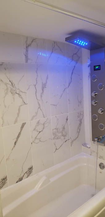 A modern bathroom with a white marble tiled shower wall, a white bathtub, and a shower panel emitting a blue LED light.