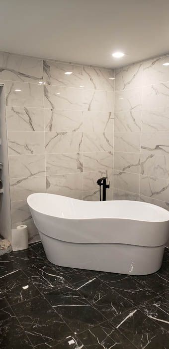A freestanding white bathtub sits against white marble-tiled walls on a black marble floor in a modern bathroom.