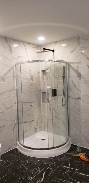 A corner shower stall with a curved glass door, black hardware, and white marble-patterned tile walls in a bathroom.