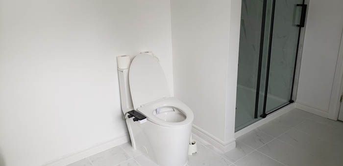 A white toilet with an attached bidet seat stands in a modern, minimalist bathroom next to a glass-walled shower.