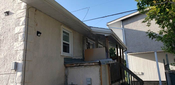 A tan stucco house exterior showing a corner, a single window, and a wooden deck structure with unfinished repairs.