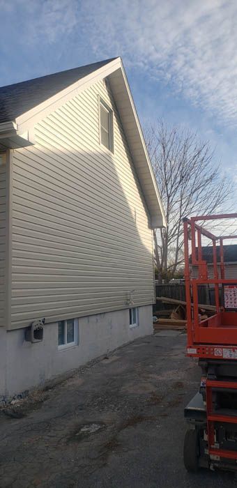 Beige vinyl siding on a gable-end wall above a concrete foundation with two small windows, next to a red scissor lift.