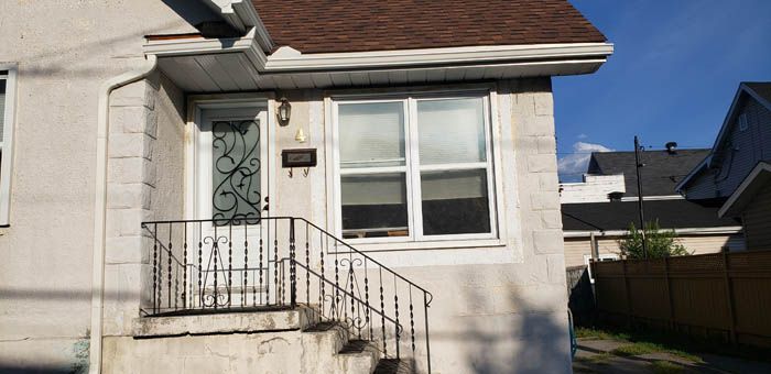 A white residential house exterior with a small porch, iron railing, storm door, and a brown shingled roof.