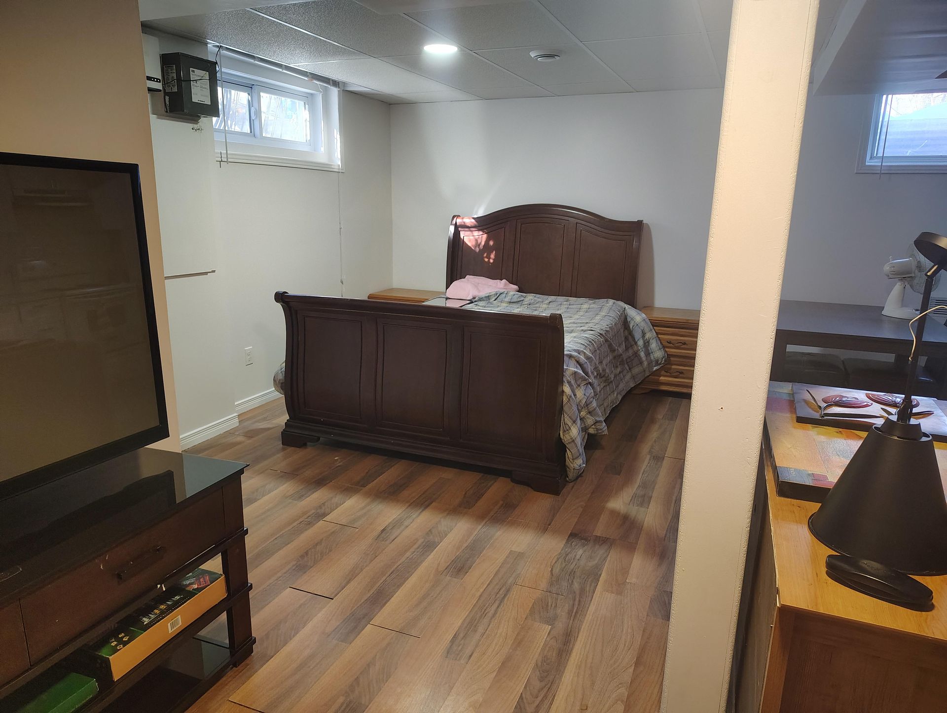 A bedroom with a dark wood bed frame, a plaid comforter, and wood-patterned flooring, viewed from an adjacent room.