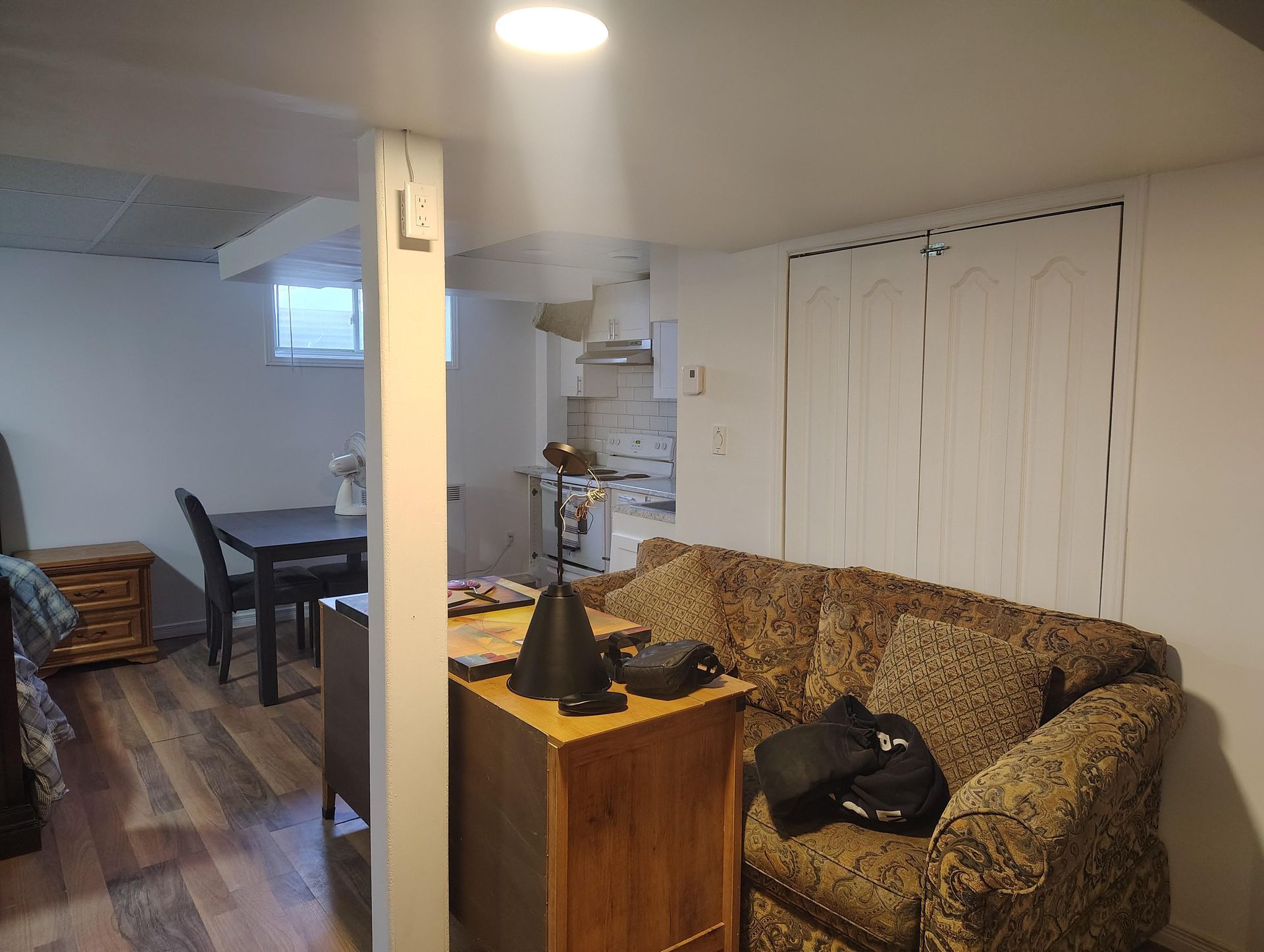 A basement interior with wood floors, a sofa, a wooden desk, a dining set, and a white support pillar.