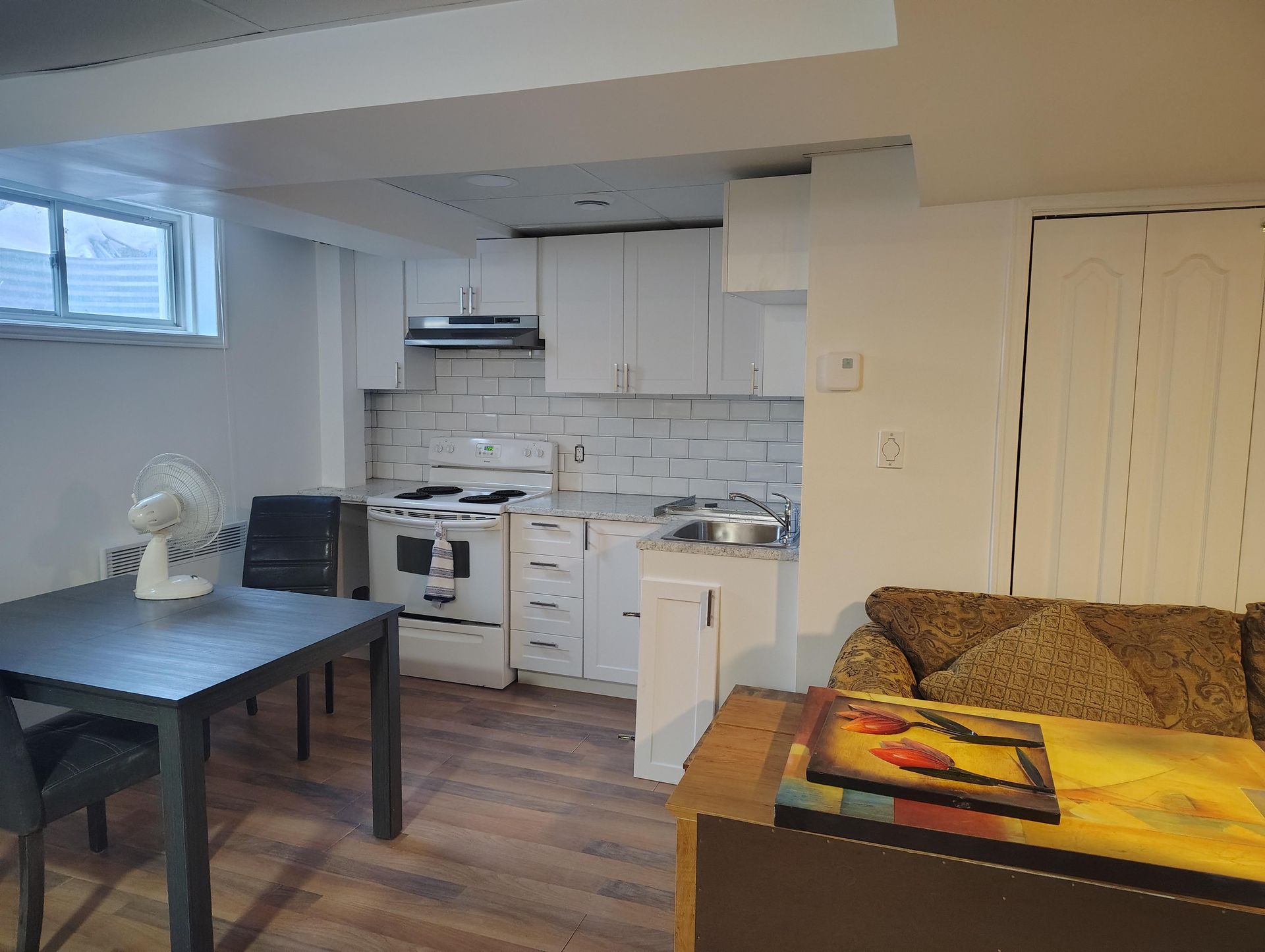 A small basement apartment featuring a white kitchen, a dining table with chairs, and a portion of a couch.