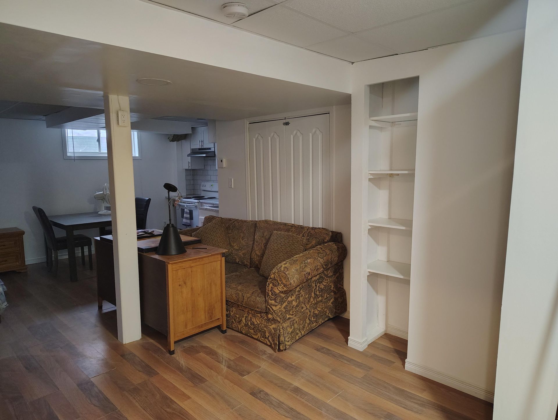 A basement living space with wood flooring, a patterned brown sofa, a desk, a dining area, and built-in white shelves.