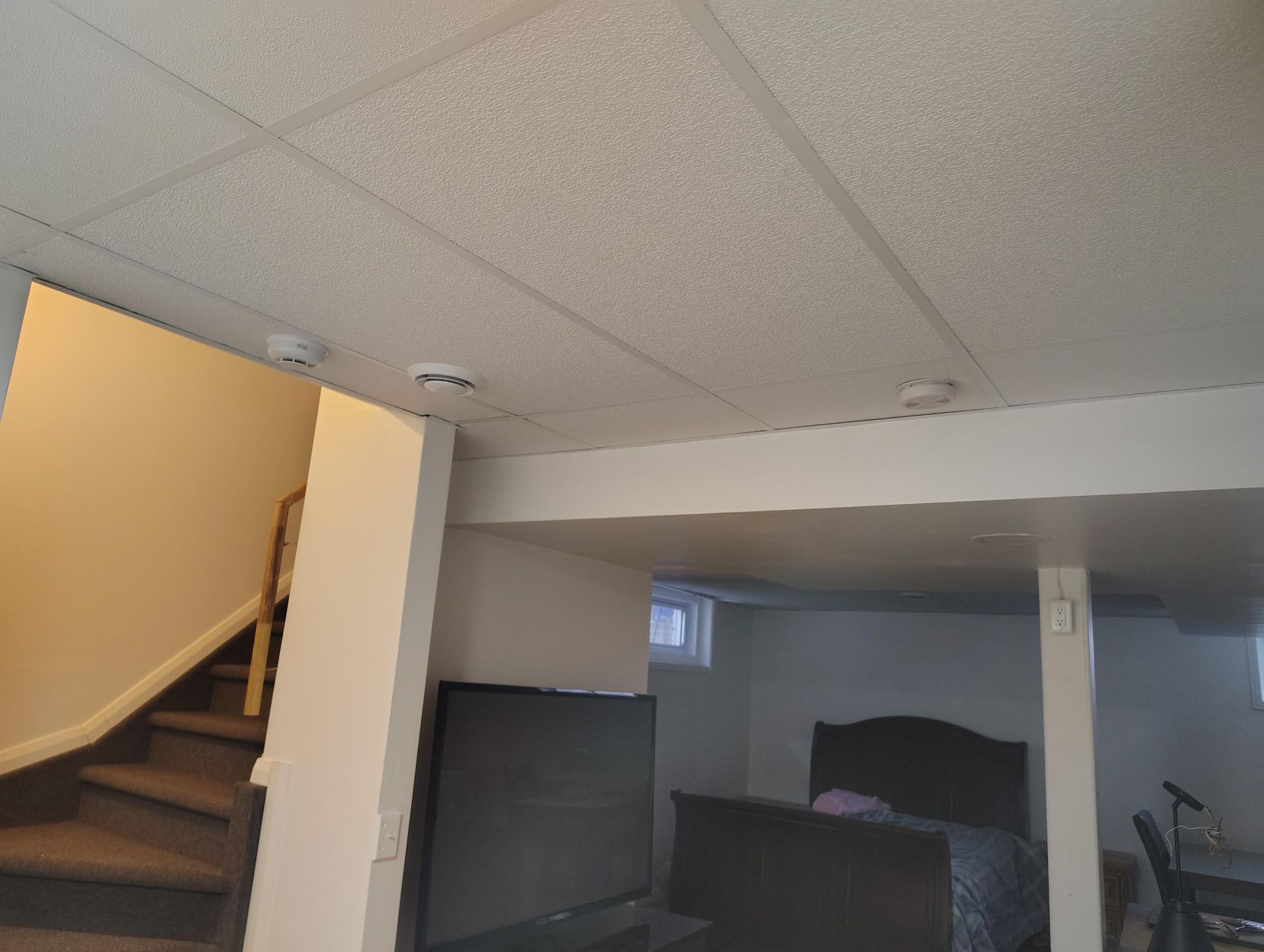 A basement interior with a carpeted staircase, a television in the foreground, and a bed in the background area.