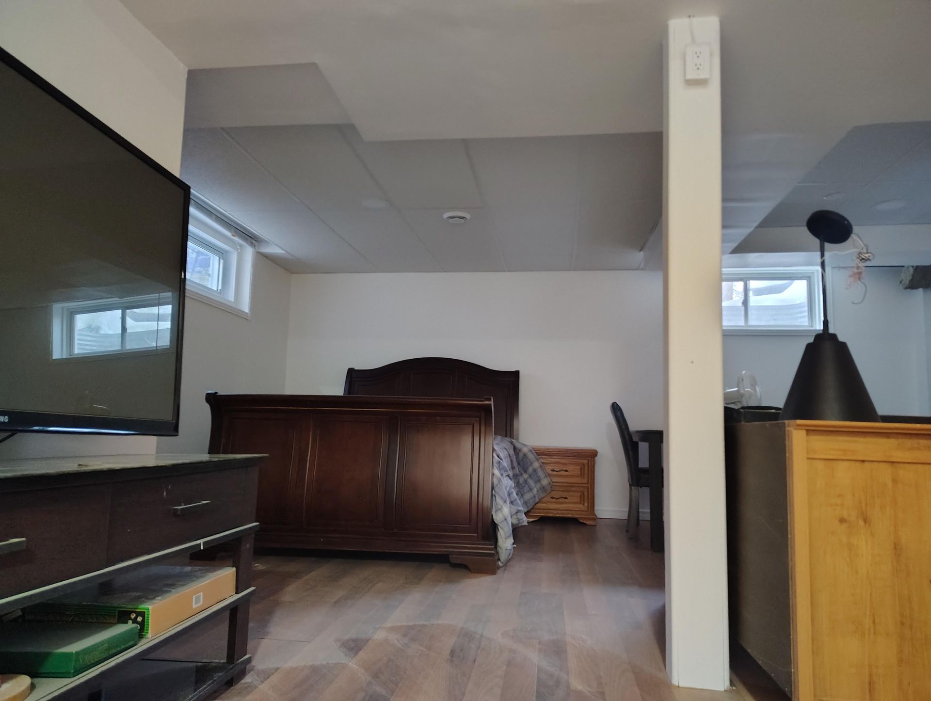 A basement bedroom with light wood flooring, a dark wooden bed frame, a TV console, a wooden nightstand, and a support column.
