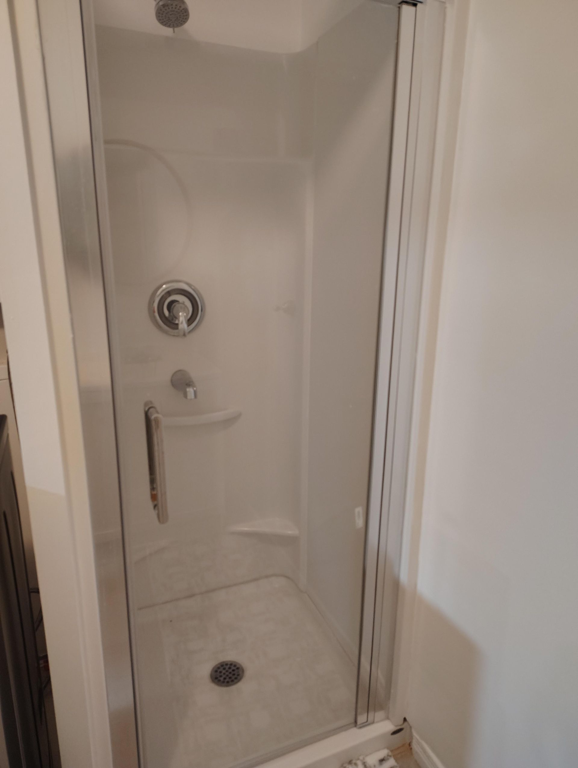 A white, walk-in shower stall with a glass door, silver faucet, overhead showerhead, and small built-in shelf.