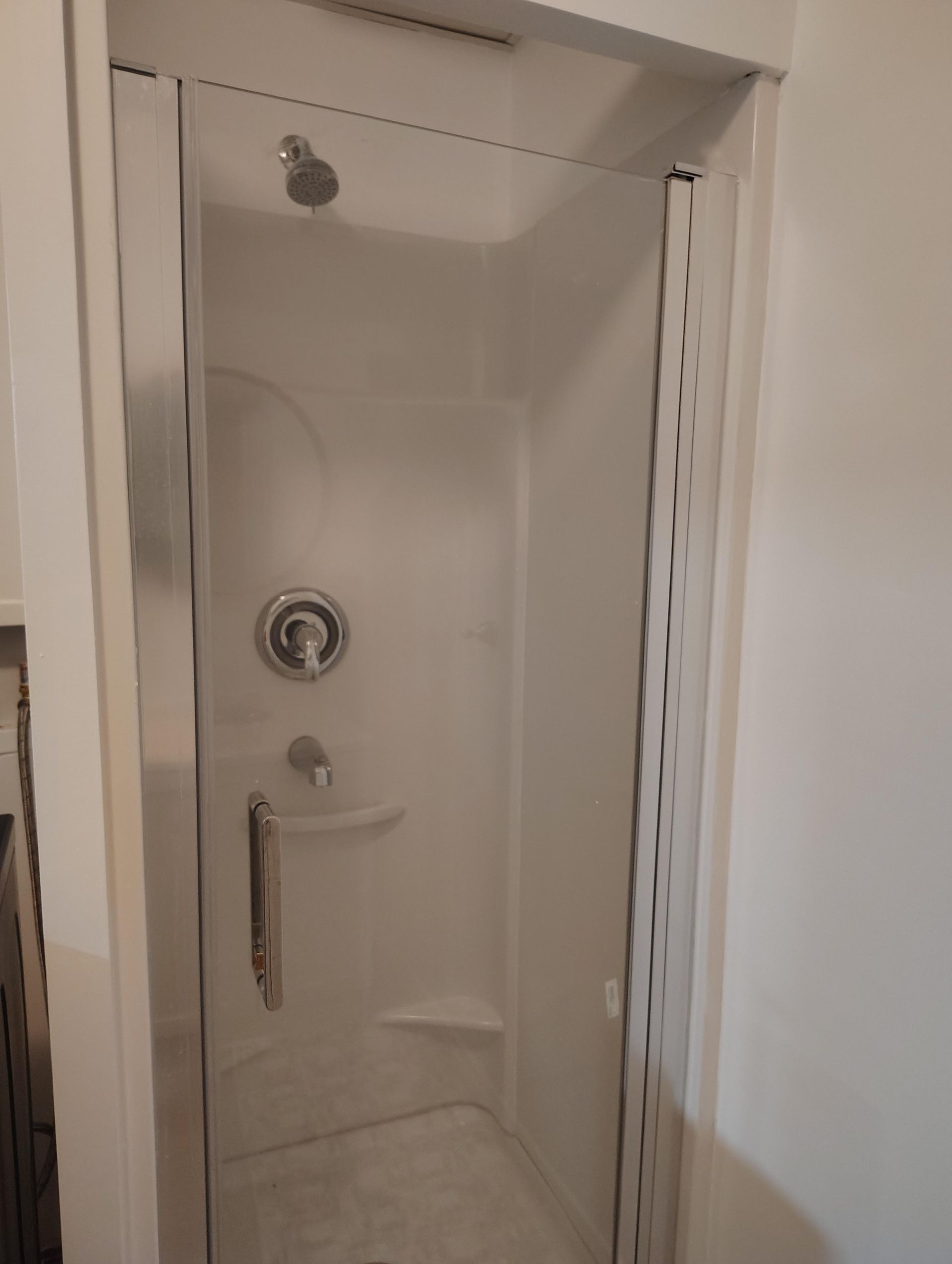 A white enclosed shower stall with a glass door, chrome fixtures, and a handle, set in a white-walled room.