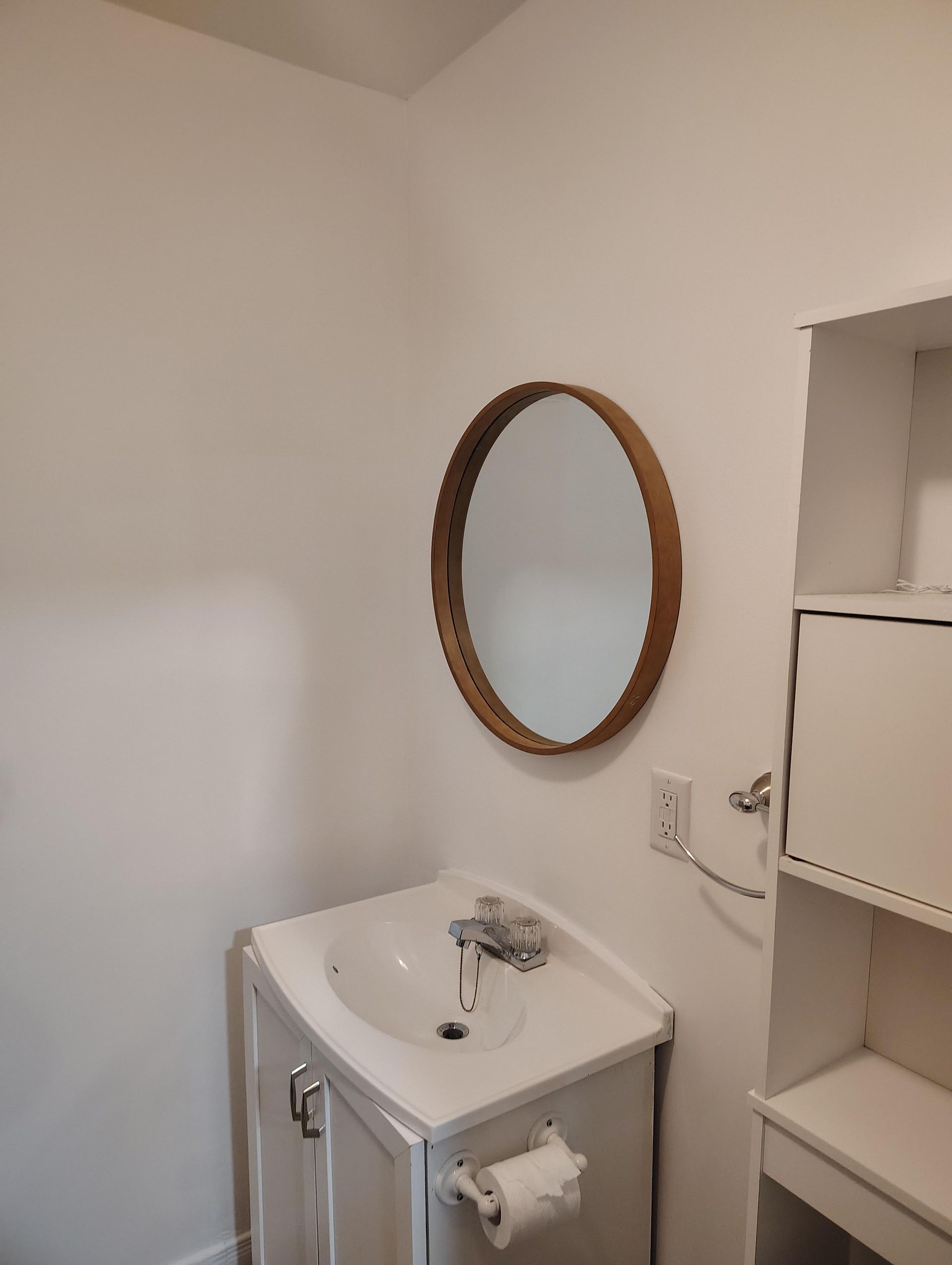 A white bathroom vanity with a chrome faucet, a wooden-framed round mirror, and an adjacent white shelving unit.