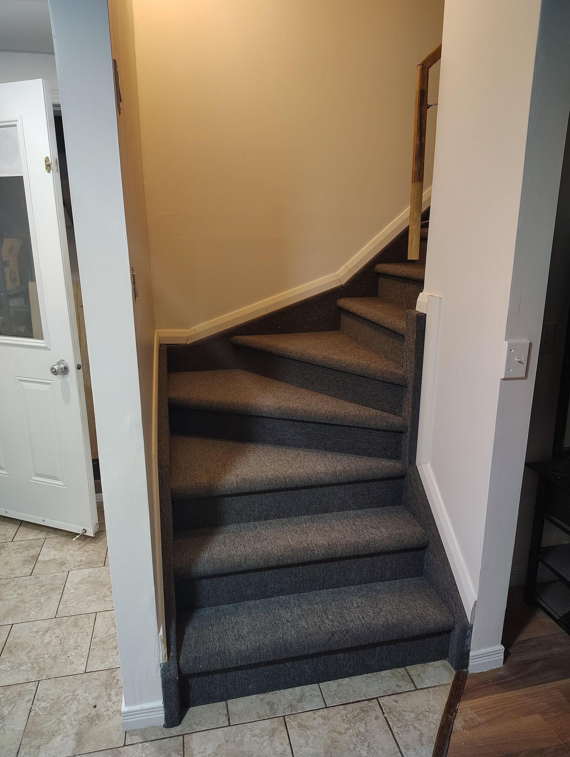 A carpeted staircase curves upward between two white walls, viewed from a tiled entryway.