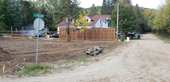 A dirt road intersection with a wooden fence, a house, trees, and a street sign on a cloudy day.