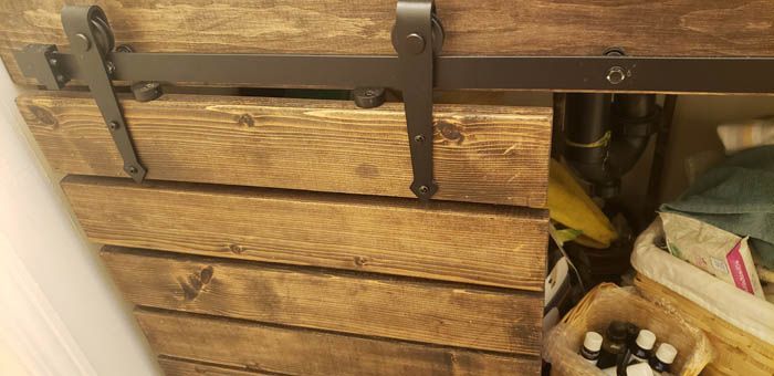 A rustic wooden sliding barn door on a metal track partially covering storage shelving under a bathroom vanity.