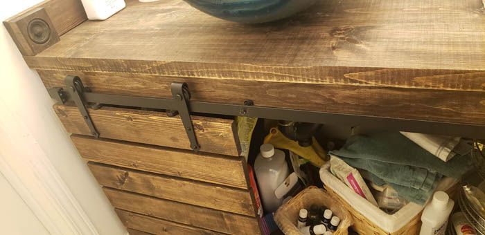 A rustic wooden bathroom vanity with a sliding barn door hardware mechanism, showing an open cabinet filled with supplies.